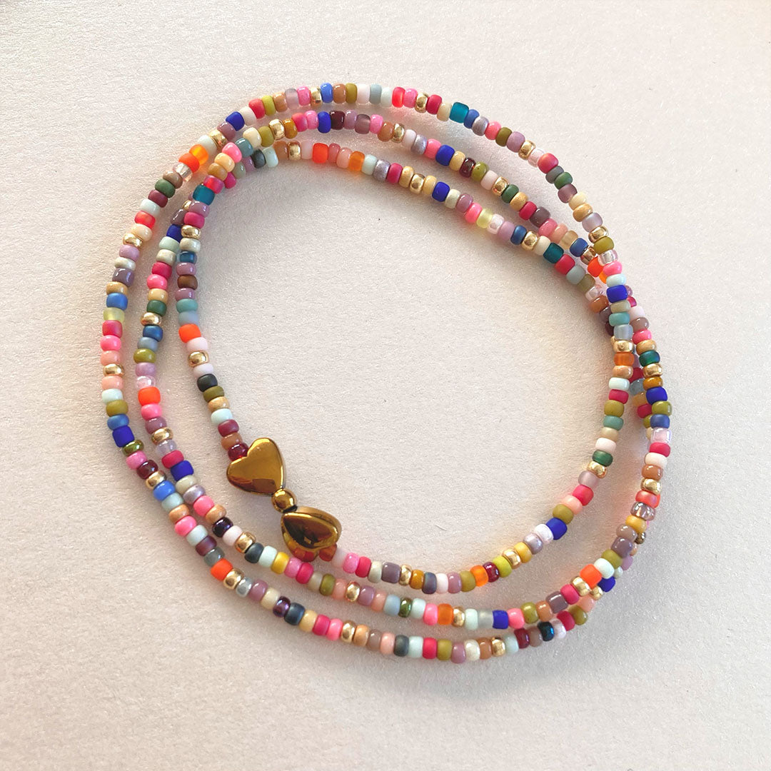 Mehrreihiges Wickelarmband aus präzisen, bunten Miyuki-Glasperlen in verschiedenen Farben wie Türkis, Pink und Gelb. In der Mitte befindet sich ein glänzender Anhänger aus zwei goldfarbenen Herzen aus Hämatit-Stein. Der Schmuck liegt auf einem neutralen, hellen Hintergrund.