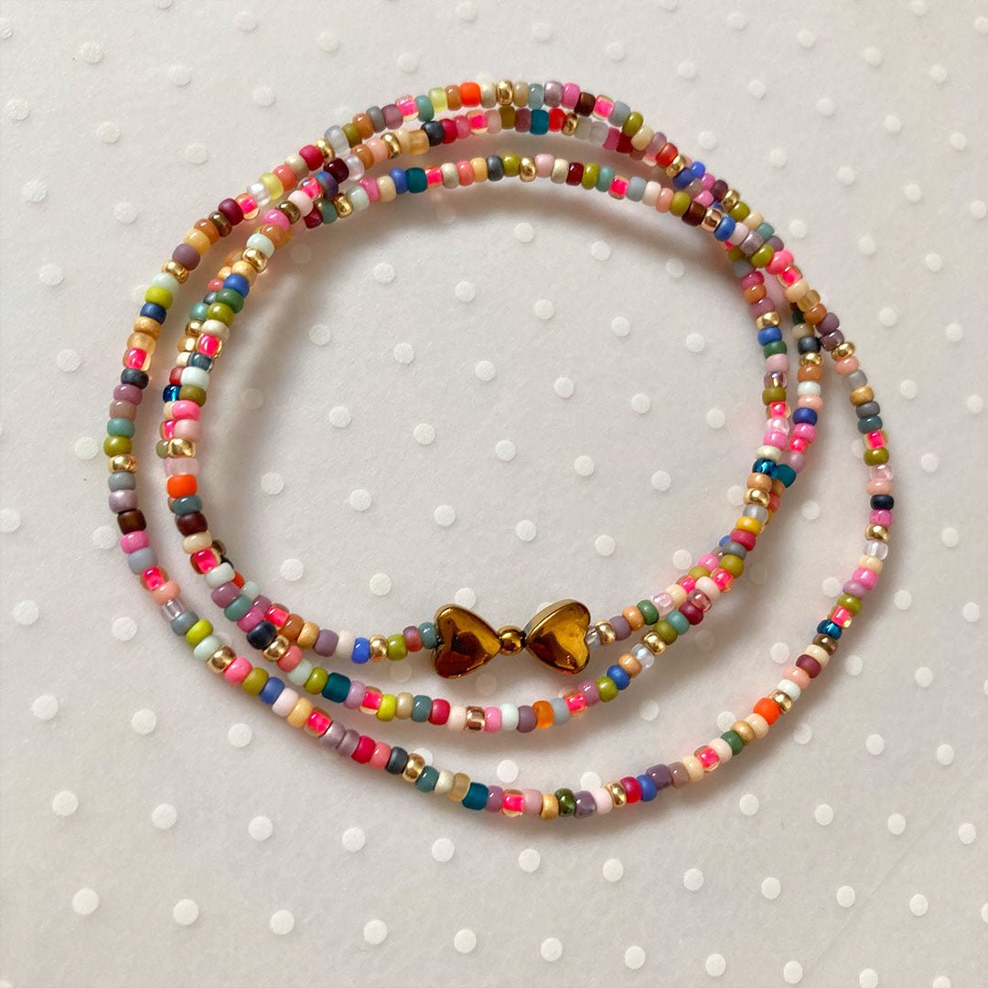 Mehrreihiges Wickelarmband aus präzisen, bunten Miyuki-Glasperlen in verschiedenen Farben wie Türkis, Pink und Gelb. In der Mitte befindet sich ein glänzender Anhänger aus zwei goldfarbenen Herzen aus Hämatit-Stein. Der Schmuck liegt auf einem neutralen, hellen Hintergrund.