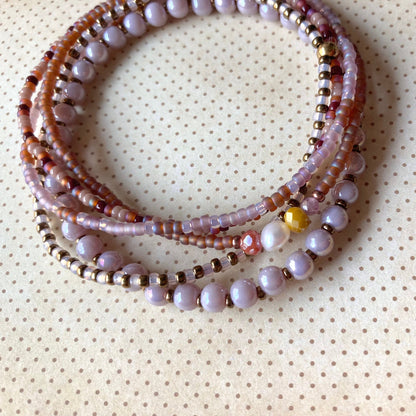 Mehrreihiges Perlenarmband in Lavendel-, Burgunder-, Braun- und Goldtönen mit unterschiedlichen Perlengrößen und -strukturen, kreisförmig angeordnet auf cremefarbenem Stoff mit Punkten. Mit metallischen Akzentperlen und bunten Steinen, darunter eine leuchtend gelbe Perle und eine Süßwasserperle.