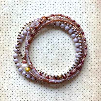 Mehrreihiges Perlenarmband in Lavendel-, Burgunder-, Braun- und Goldtönen mit unterschiedlichen Perlengrößen und -strukturen, kreisförmig angeordnet auf cremefarbenem Stoff mit Punkten. Mit metallischen Akzentperlen und bunten Steinen, darunter eine leuchtend gelbe Perle und eine Süßwasserperle.