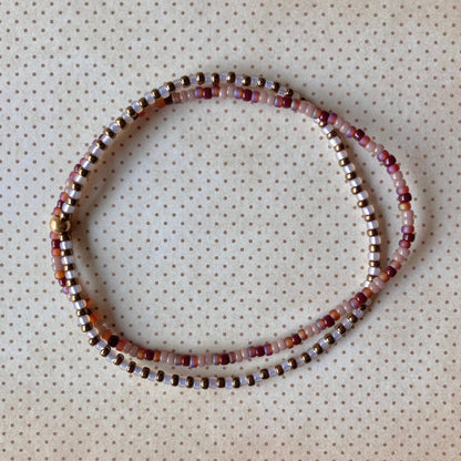 Zweireihiges Perlenarmband in Lavendel-, Burgunder-, Braun- und Goldtönen, kreisförmig angeordnet auf cremefarbenem Stoff mit Punkten.