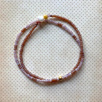 Zweireihiges Perlenarmband in Lavendel-, Burgunder-, Braun- und Goldtönen mit unterschiedlichen Perlengrößen und -strukturen, kreisförmig angeordnet auf cremefarbenem Stoff mit Punkten. Mit metallischen Akzentperlen und bunten Steinen, darunter eine leuchtend gelbe Perle und eine Süßwasserperle.