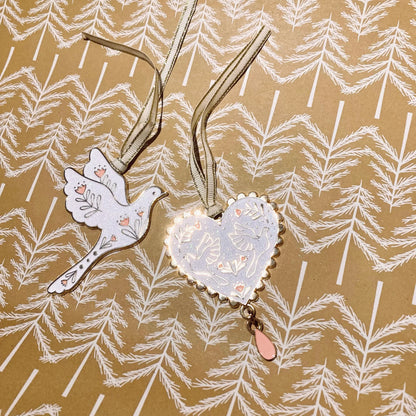 Zwei weiße Anhänger mit goldfarbenen Rändern und floralen Mustern, einer in Taubenform, der andere herzförmig mit einem Tropfenanhänger, auf braunem Papier mit weißem Tannenzweigmuster.