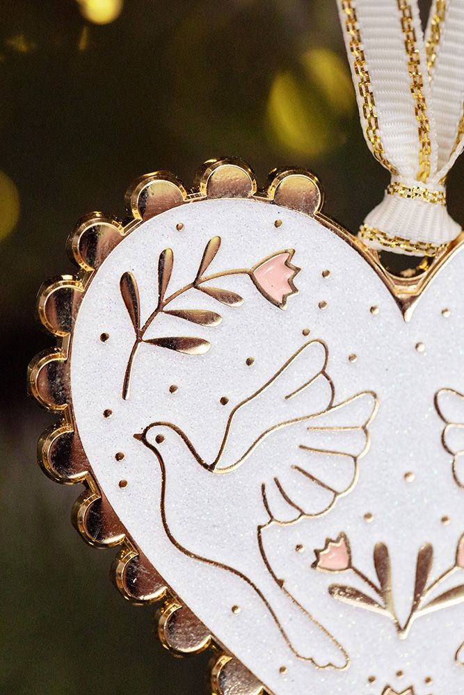 Herzförmiger Christbaumschmuck aus weißem Material mit goldfarbenem Rand, verziert mit zwei Tauben und floralen Mustern, hängt an einem Tannenzweig.