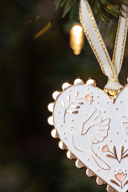 Herzförmiger Christbaumschmuck aus weißem Material mit goldfarbenem Rand, verziert mit zwei Tauben und floralen Mustern, hängt an einem Tannenzweig.