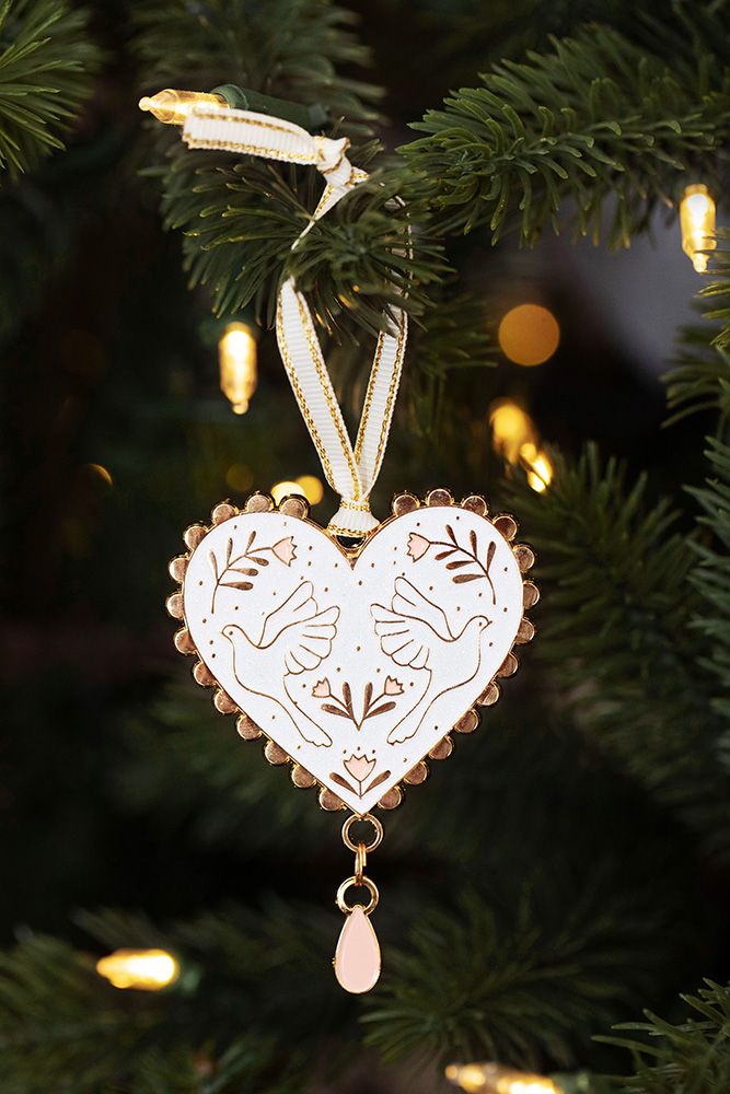 Herzförmiger Christbaumschmuck aus weißem Material mit goldfarbenem Rand, verziert mit zwei Tauben und floralen Mustern, hängt an einem Tannenzweig.