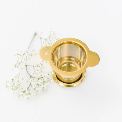 Goldfarbener Teesieb aus Metall mit zwei Griffen, daneben weiße Blütenzweige.