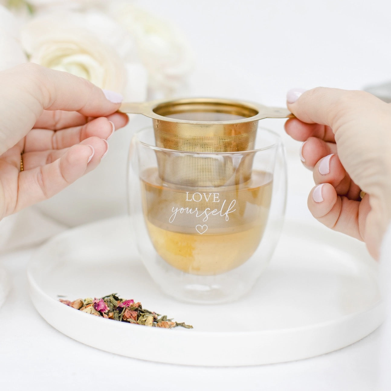 Hände halten einen goldfarbenen Teesieb über eine doppelwandige Glastasse mit der Aufschrift 'LOVE yourself' auf einem weißen Tablett mit losem Tee.