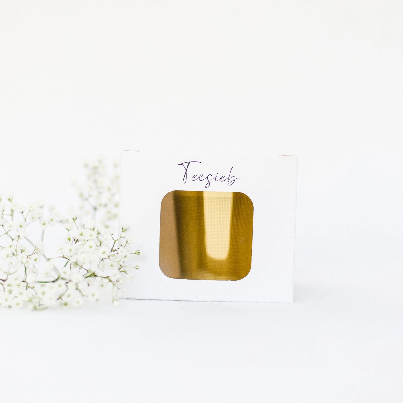 Weißer Karton mit Sichtfenster, darin goldfarbenes Teesieb, links weiße Blütenzweige.