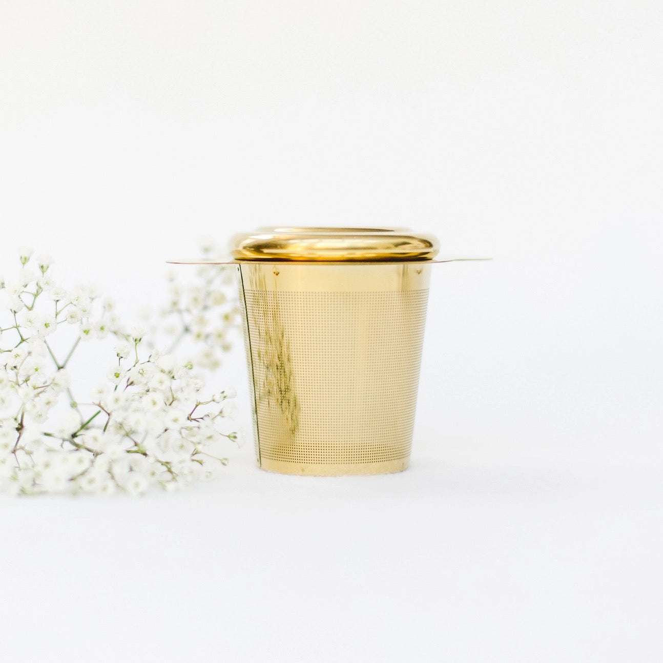 Goldfarbener Teesieb mit Deckel, daneben weiße Blütenzweige, auf hellem Untergrund.