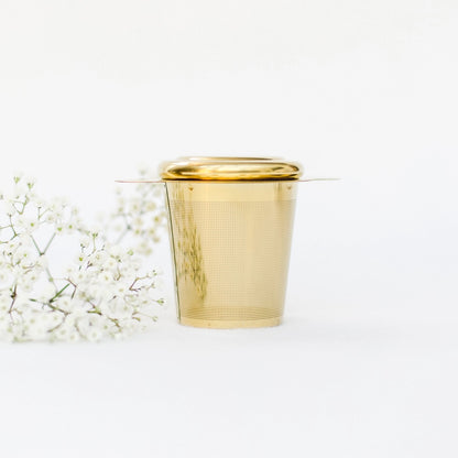 Goldfarbener Teesieb mit Deckel, daneben weiße Blütenzweige, auf hellem Untergrund.