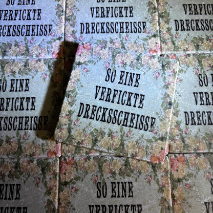 Quadratische Fliesen  mit Blumenmuster und dem Text 'SO EINE VERFICKTE DRECKSSCHEISSE' in Großbuchstaben