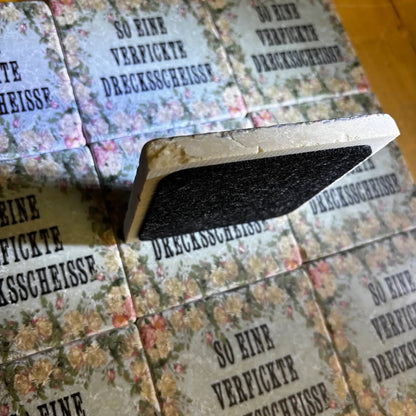 Quadratische Fliesen  mit Blumenmuster und dem Text 'SO EINE VERFICKTE DRECKSSCHEISSE' in Großbuchstaben , eine Fliese mit Filz von hinten