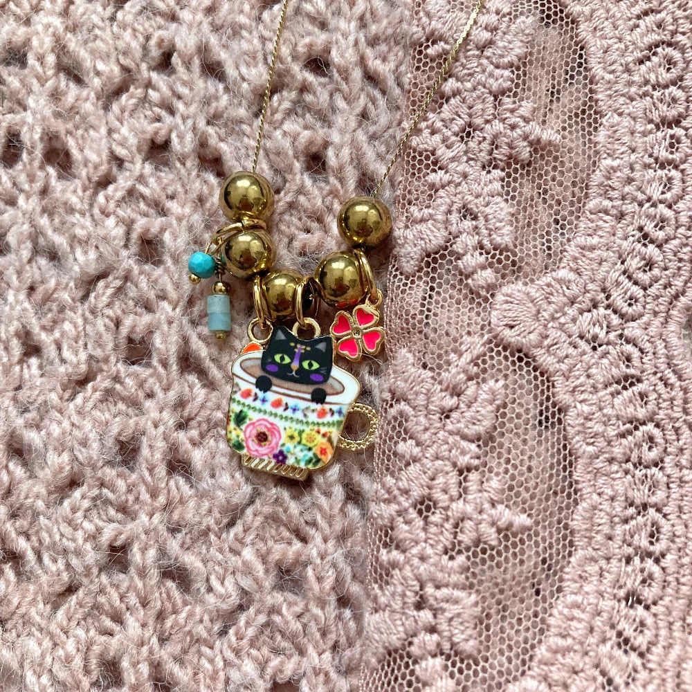 Halskette mit goldfarbenen Kugeln und Anhängern, darunter eine Tasse mit schwarzer Katze, ein rotes Herz und ein vierblättriges Kleeblatt.