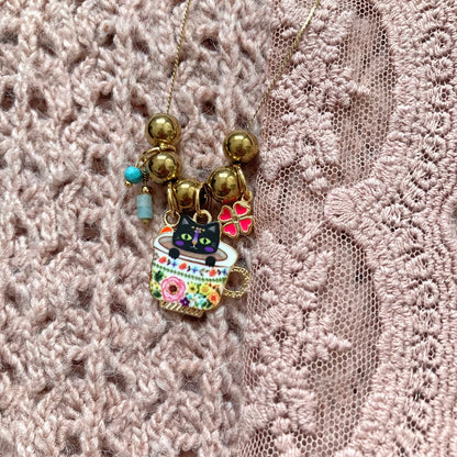 Halskette mit goldfarbenen Kugeln und Anhängern, darunter eine Tasse mit schwarzer Katze, ein rotes Herz und ein vierblättriges Kleeblatt.