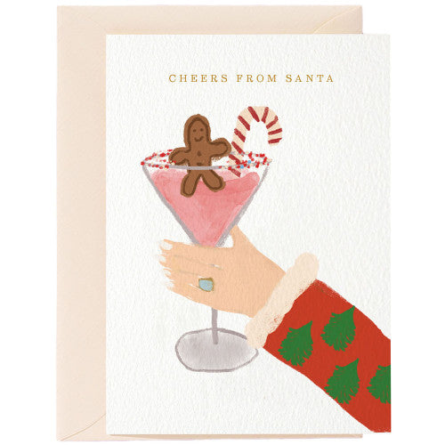 Hand in rotem Pullover mit grünen Tannenzweigen hält Cocktailglas mit rosa Getränk, Zuckerstange und Lebkuchenmann.