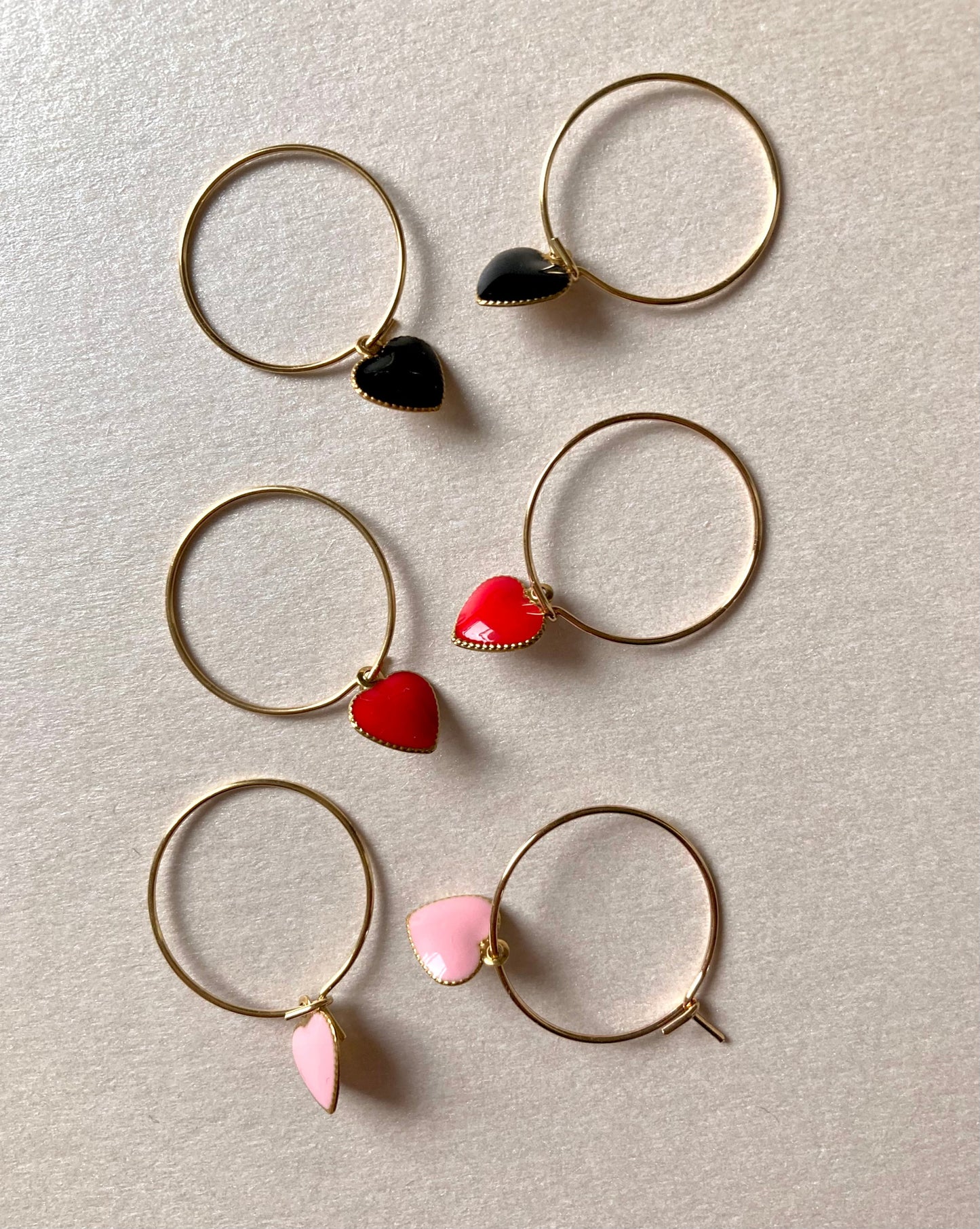 3 Paar goldfarbene Creolen mit herzförmigen Anhängern in Schwarz, Rot und Rosa, auf hellem Untergrund.