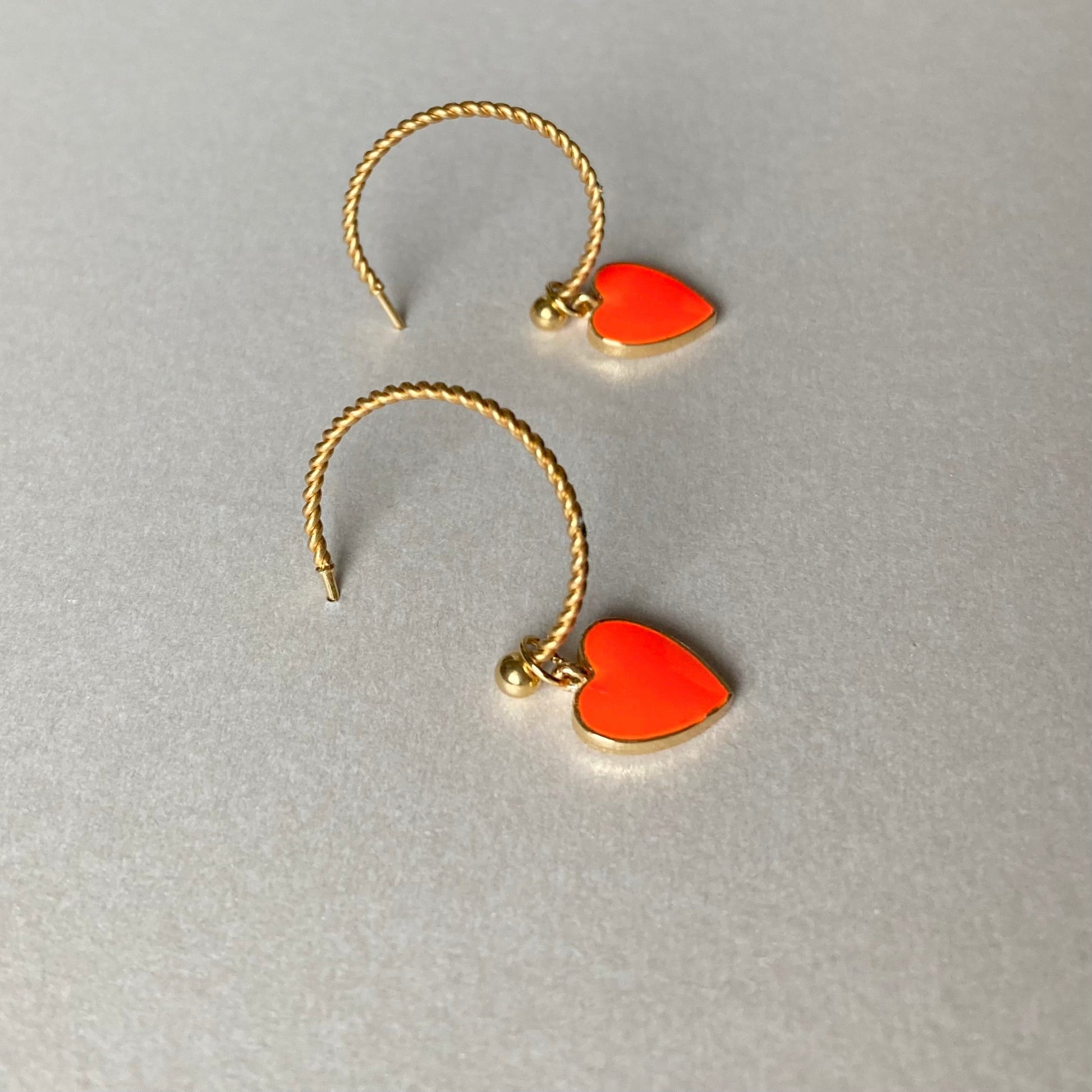 Zwei goldfarbene, gedrehte Creolen mit je einem neonorangen Herzanhänger auf grauem Untergrund.
