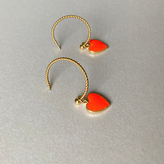 Zwei goldfarbene, gedrehte Creolen mit je einem neonorangen Herzanhänger auf grauem Untergrund.