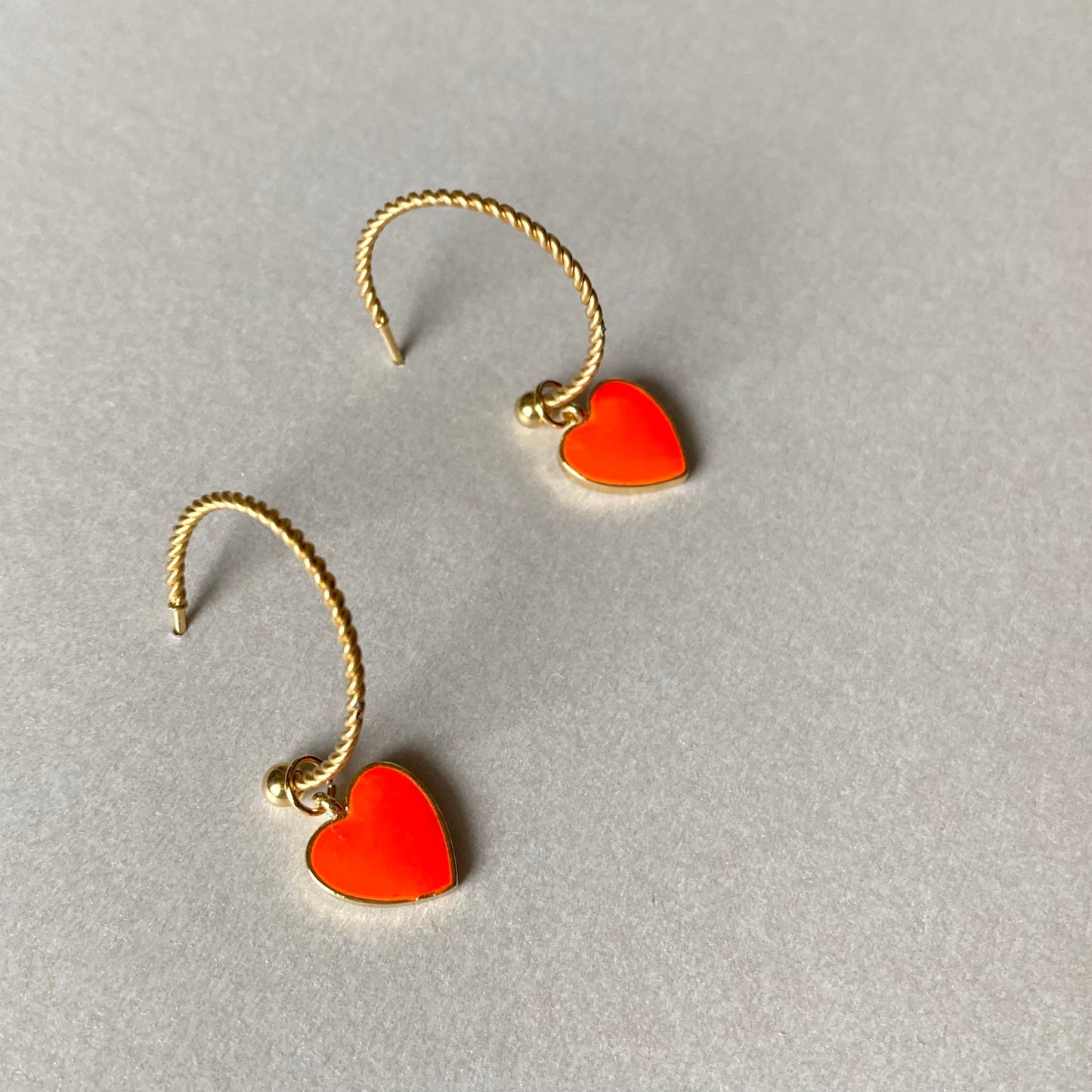 Zwei goldfarbene, gedrehte Creolen mit je einem neonorangen Herzanhänger auf grauem Untergrund.