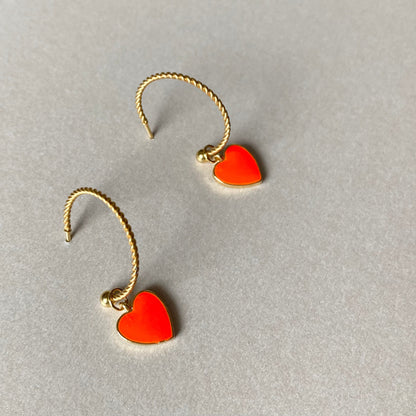 Zwei goldfarbene, gedrehte Creolen mit je einem neonorangen Herzanhänger auf grauem Untergrund.