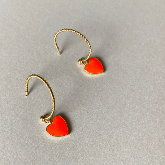 Zwei goldfarbene, gedrehte Creolen mit je einem neonorangen Herzanhänger auf grauem Untergrund.