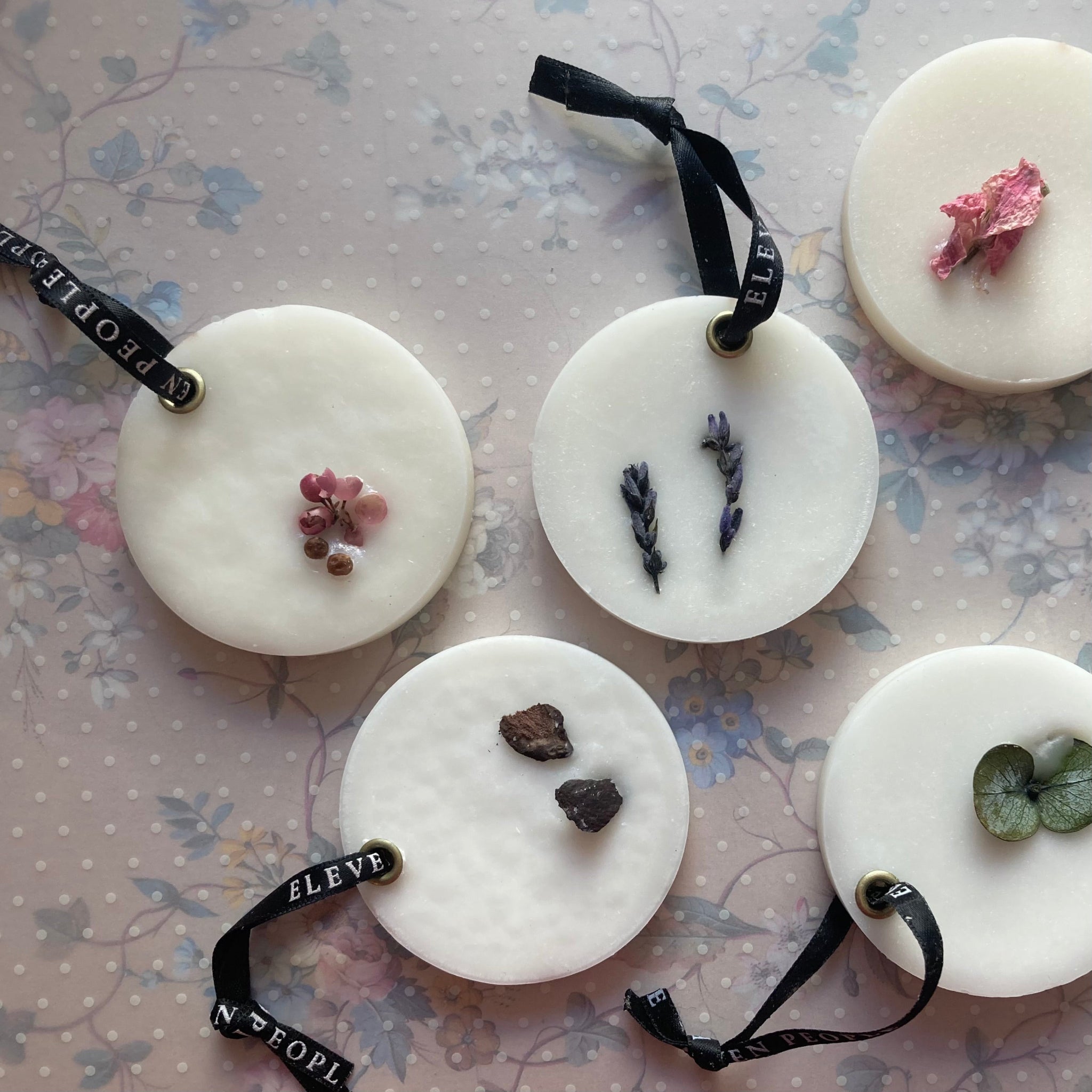 Fünf weiße runde Wachsscheiben mit getrockneten Blüten und Kräutern, jede mit schwarzem Band zum Aufhängen