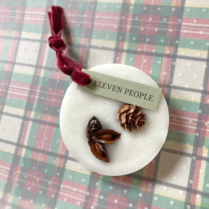 Ein runder, weißer Sojawachs-Lufterfrischer, verziert mit pflanzlichen Elementen. Der Lufterfrischer hat ein burgunderfarbenes Samtband zum Aufhängen und liegt auf einem festlich karierten Hintergrund in gedeckten Weihnachtsfarben Grün, Rot und Creme mit weißen Punkten.