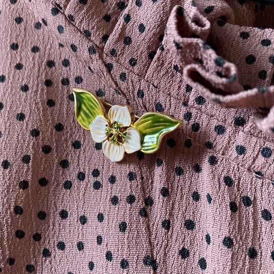 Goldfarbene Brosche in Form einer Blüte mit weißen Blütenblättern und zwei grünen Blättern, verziert mit kleinen grünen Punkten in der Mitte an einem gepunkteten Stoff.