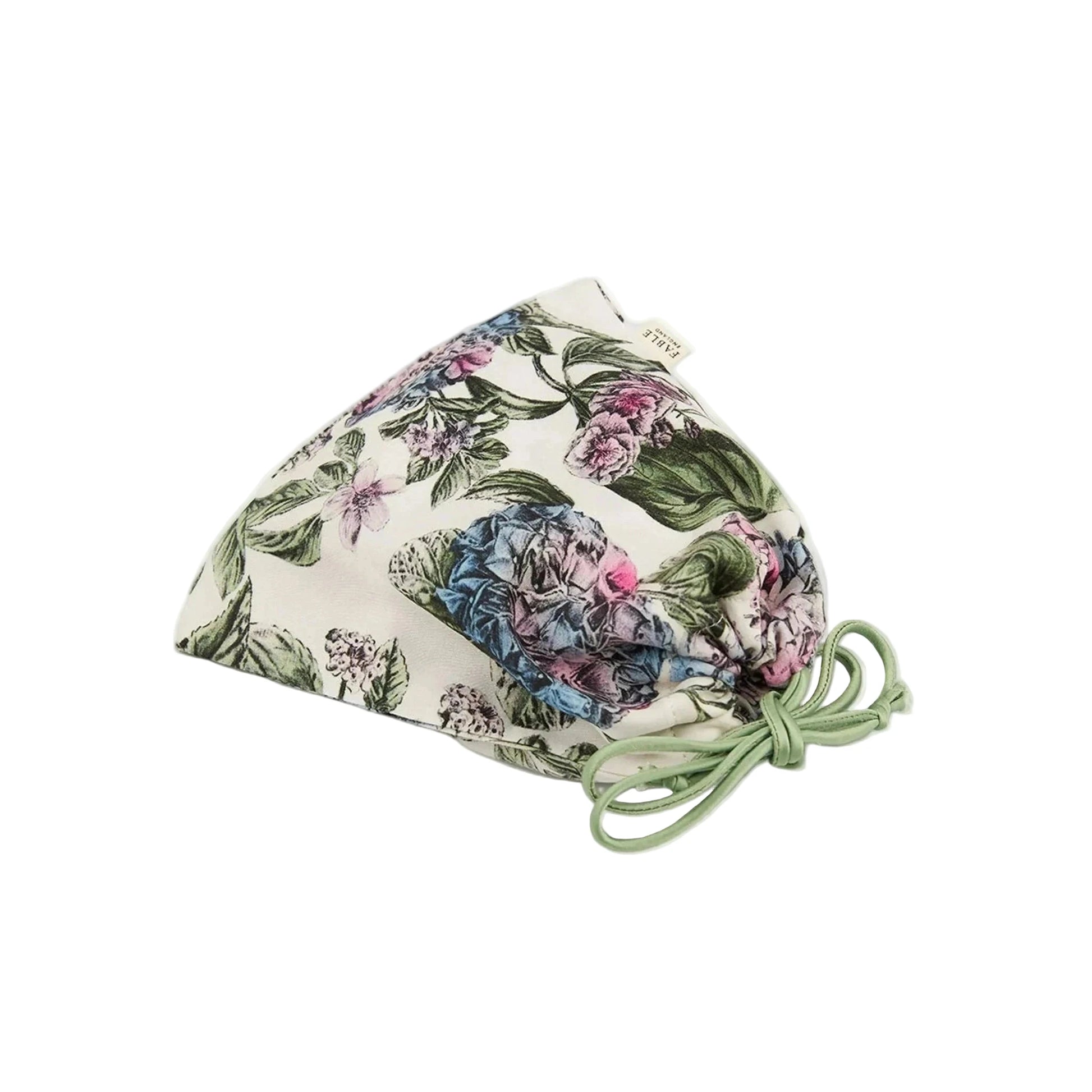 Zusammengelegte Schlafmaske mit botanischem Print in einem passenden Aufbewahrungsbeutel aus demselben Material, Beutel mit Kordelzug in Dunkelblau zum Zuziehen, Marken-Etikett sichtbar, zeigt die platzsparende Aufbewahrungslösung für unterwegs.