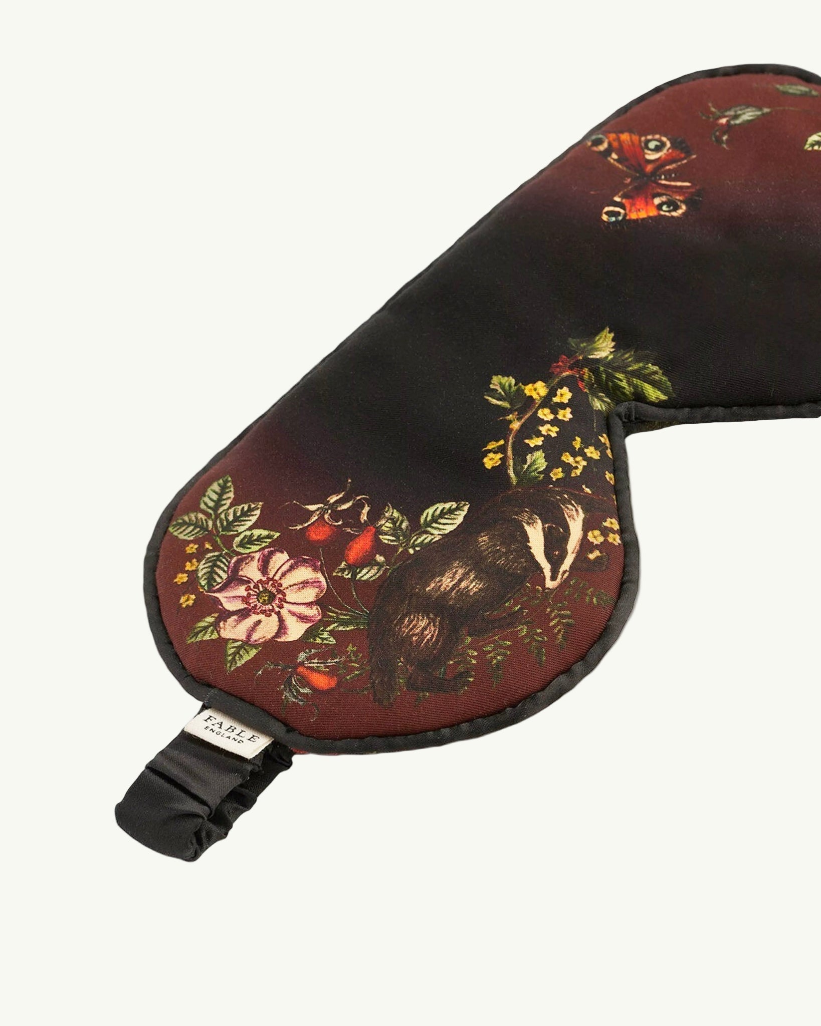 Schlafmaske mit künstlerischem Naturmotiv auf rotbraunem und schwarzem Untergrund: detaillierter Igel, Tagpfauenauge-Schmetterling, Wildblumen, Farnblätter und Beeren in warmen Herbstfarben wie Rostrot, Gelb, Grün und Orange, schwarzes Satinband mit Etikett, naturalistischer Vintage-Stil
