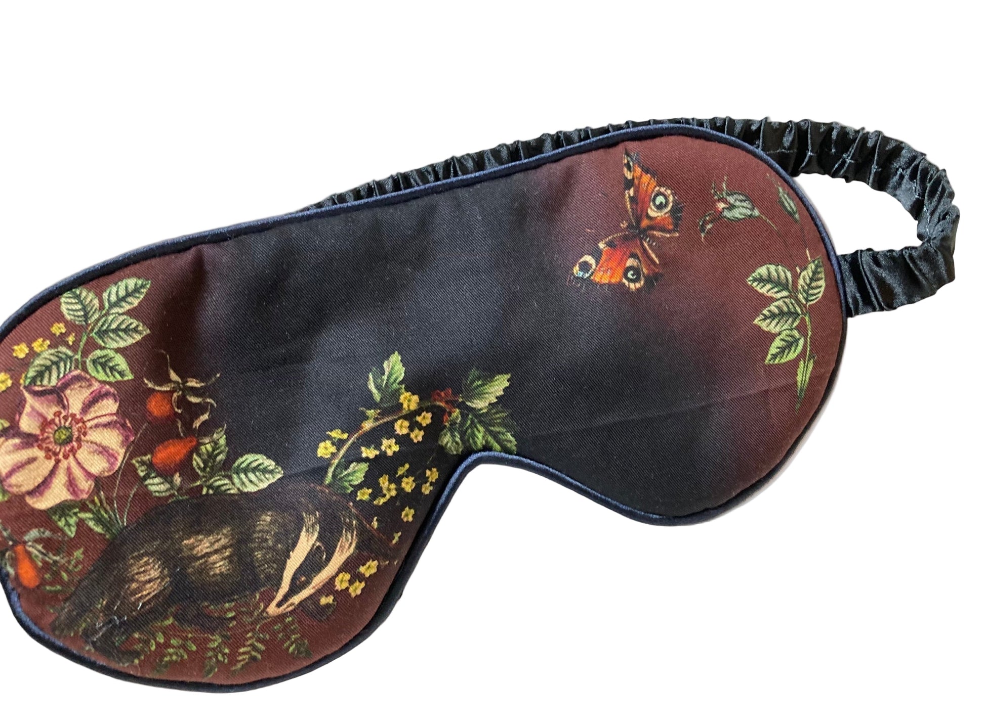 Schlafmaske mit künstlerischem Naturmotiv auf rotbraunem und schwarzem Untergrund: detaillierter Igel, Tagpfauenauge-Schmetterling, Wildblumen, Farnblätter und Beeren in warmen Herbstfarben wie Rostrot, Gelb, Grün und Orange, schwarzes Satinband mit Etikett, naturalistischer Vintage-Stil