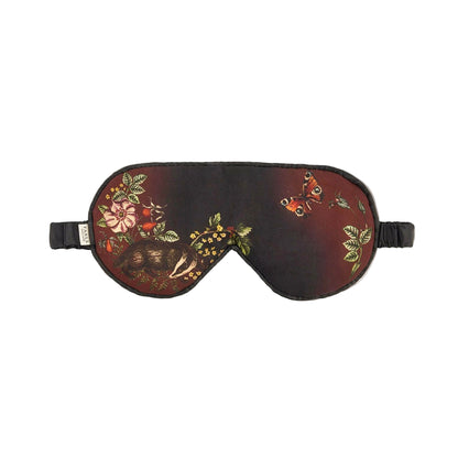 Schlafmaske mit künstlerischem Naturmotiv auf rotbraunem und schwarzem Untergrund: detaillierter Igel, Tagpfauenauge-Schmetterling, Wildblumen, Farnblätter und Beeren in warmen Herbstfarben wie Rostrot, Gelb, Grün und Orange, schwarzes Satinband mit Etikett, naturalistischer Vintage-Stil