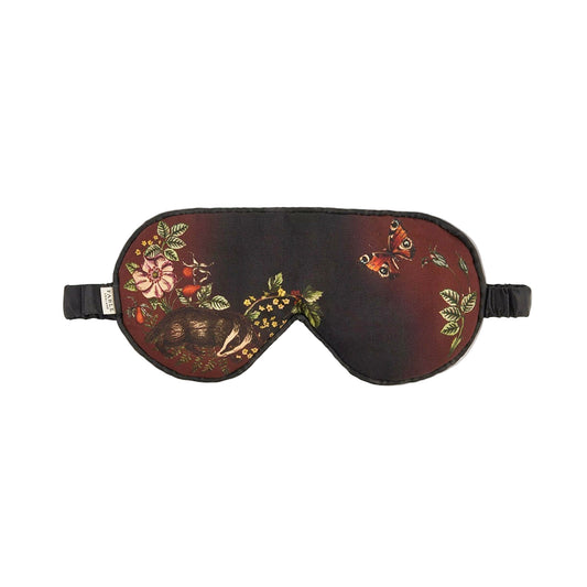Schlafmaske mit künstlerischem Naturmotiv auf rotbraunem und schwarzem Untergrund: detaillierter Igel, Tagpfauenauge-Schmetterling, Wildblumen, Farnblätter und Beeren in warmen Herbstfarben wie Rostrot, Gelb, Grün und Orange, schwarzes Satinband mit Etikett, naturalistischer Vintage-Stil