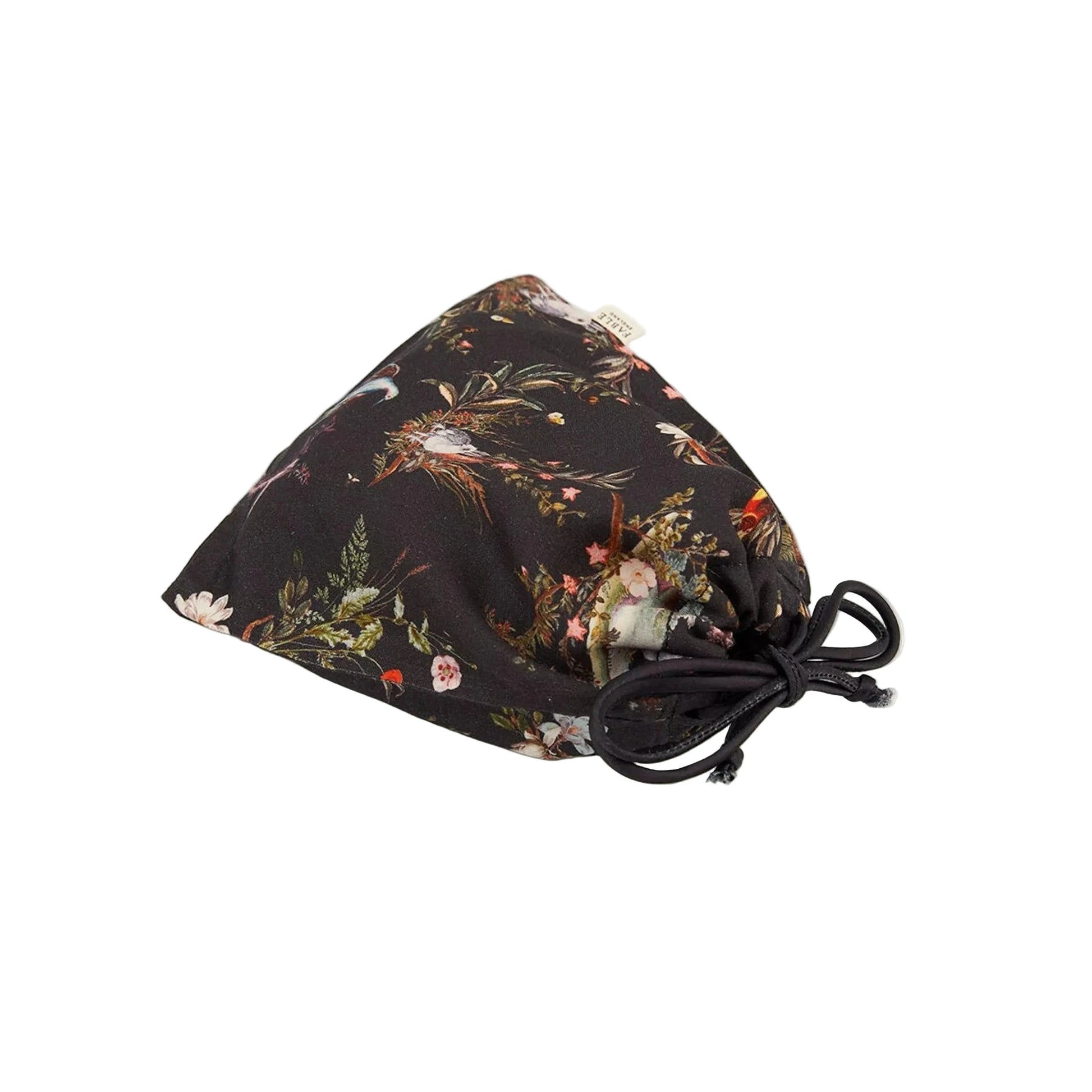 Zusammengelegte Schlafmaske mit botanischem Print in einem passenden Aufbewahrungsbeutel aus demselben Material, Beutel mit Kordelzug in Dunkelblau zum Zuziehen, Marken-Etikett sichtbar, zeigt die platzsparende Aufbewahrungslösung für unterwegs