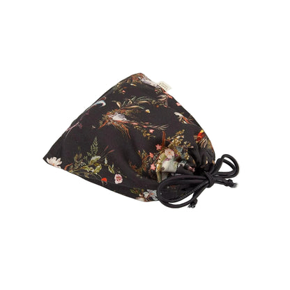 Zusammengelegte Schlafmaske mit botanischem Print in einem passenden Aufbewahrungsbeutel aus demselben Material, Beutel mit Kordelzug in Dunkelblau zum Zuziehen, Marken-Etikett sichtbar, zeigt die platzsparende Aufbewahrungslösung für unterwegs