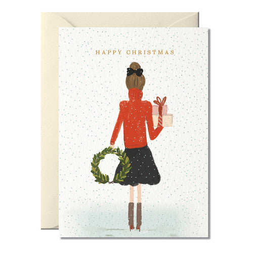 Weihnachtskarte mit Illustration einer Frau von hinten, die einen grünen Kranz in der linken Hand und ein Geschenk in der rechten Hand hält, darüber der Text 'HAPPY CHRISTMAS'.