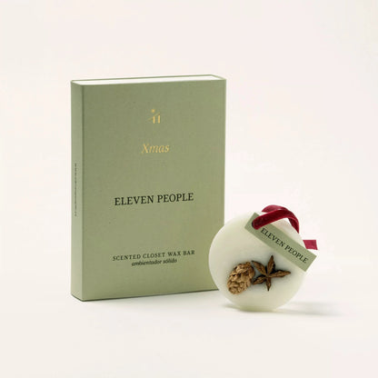 Der weihnachtlich duftende Wachsriegel „ELEVEN PEOPLE“ für den Kleiderschrank wird in einer salbeigrünen Verpackungsbox präsentiert. Die Box trägt oben den goldenen Text „11 Xmas“ und darunter „ELEVEN PEOPLE SCENTED CLOSET WAX BAR ambientador sólido“. Daneben befindet sich der runde weiße Sojawachs-Lufterfrischer, verziert mit natürlichen Elementen wie einem Tannenzapfen und Sternanis. Auf der Oberfläche ist „ELEVEN PEOPLE“ eingeprägt und an einer burgunderfarbenen Samtschleife befestigt.