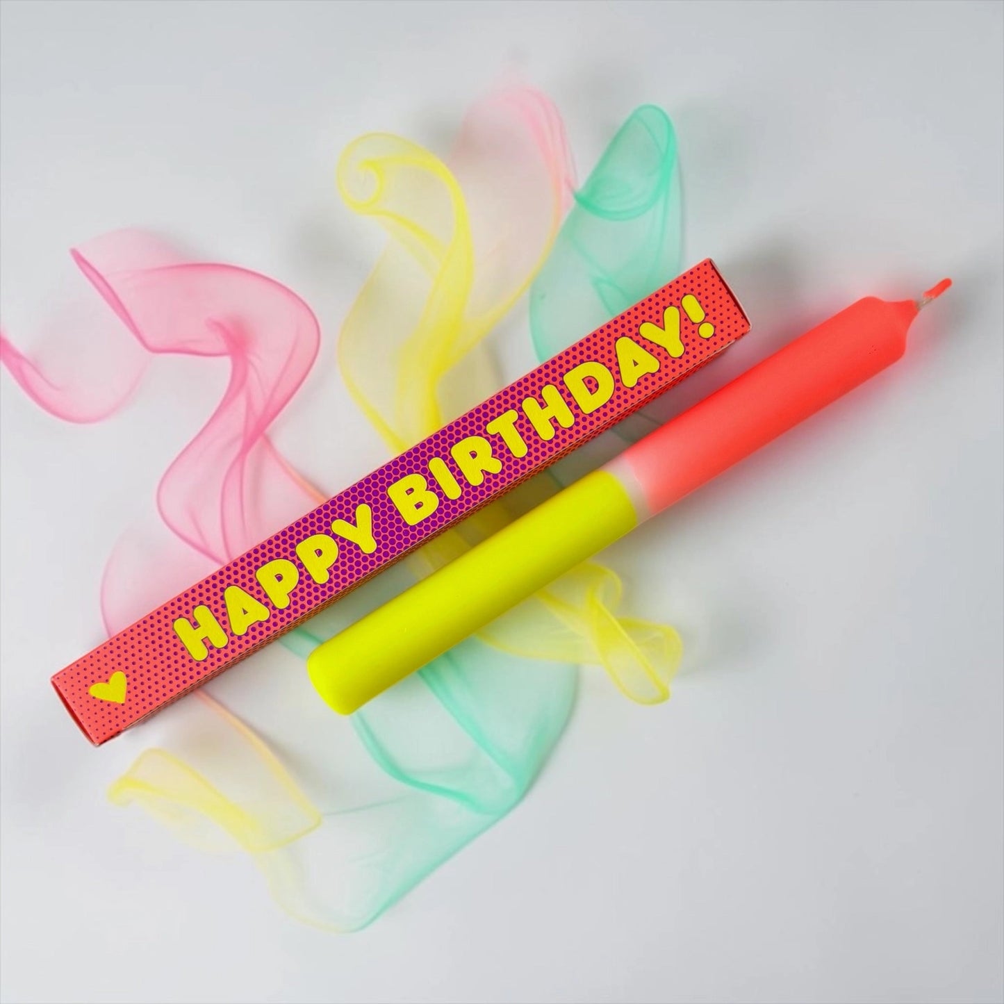 Zweifarbige Stabkerze in Neon-Gelb und Neon-Orange auf weißem Hintergrund mit bunten Chiffonbändern. Daneben die rechteckige Verpackung in Orange mit Punktmuster und dem gelben Aufdruck „HAPPY BIRTHDAY!“.