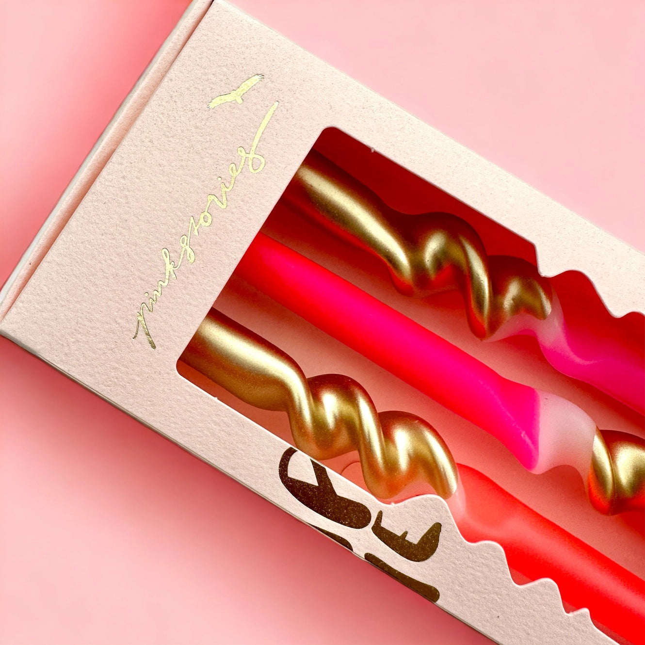 Verpackung mit drei Kerzen, zwei pink und eine rot, jeweils mit goldfarbenem Spiraldesign, vor rosa Hintergrund.