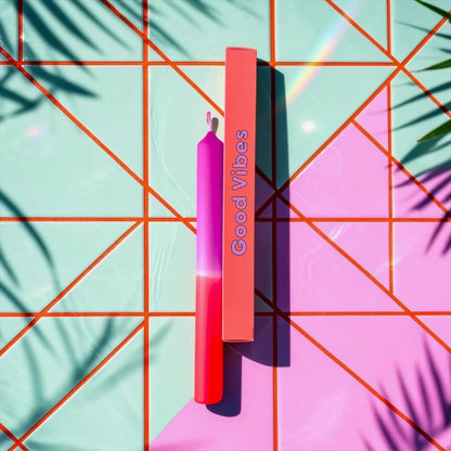 Eine Dip-Dye-Stabkerze von Pink Stories mit einem Farbverlauf von Pfirsich-Orange zu kräftigem Beere, neben der gleichfarbigen Geschenkbox mit der Aufschrift „Good Vibes“.