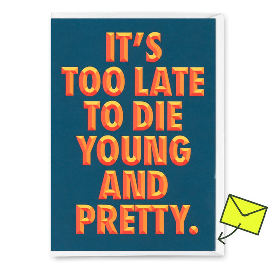 Eine Geburtstagskarte mit dunklem blaugrünem/marineblauem Hintergrund und fettem, dreidimensionalem Text in Orange und Gelb: „It’s too late to die young and pretty.“ Die Buchstaben erzeugen einen Retro-3D-Effekt mit Schatten. Oben rechts befindet sich ein gelber „Deli Cards“-Aufkleber. 