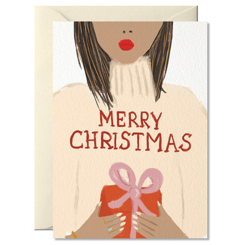 Illustration einer Frau mit braunen Haaren und rotem Lippenstift, die einen roten Geschenk mit rosa Schleife hält. Text 'MERRY CHRISTMAS' auf cremefarbenem Pullover.