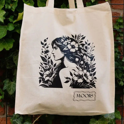 Beige Stofftasche mit schwarzem Druck einer Frau mit langem Haar, umgeben von Blumen und Blättern, darunter Schriftzug 'MOOIS'