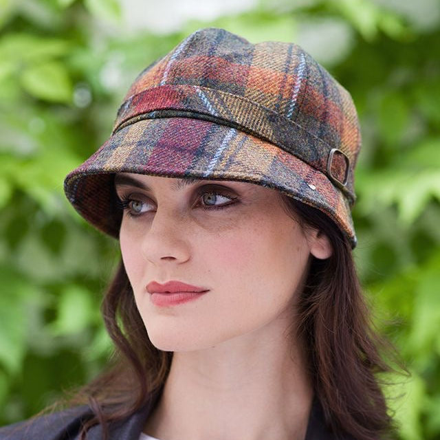 Eine Frau mit braun orangefarbenem, kariertem Flapper Hut, weißem Oberteil und schwarzem Blazer vor grünem Laub im Hintergrund.