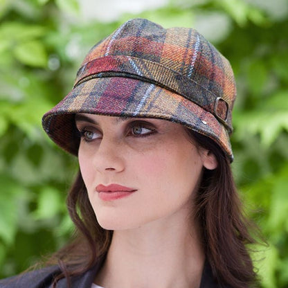 Eine Frau mit braun orangefarbenem, kariertem Flapper Hut, weißem Oberteil und schwarzem Blazer vor grünem Laub im Hintergrund.