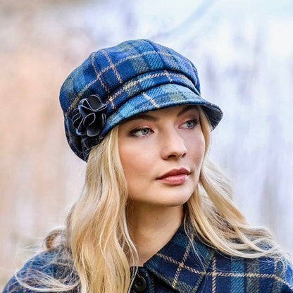 Blonde Frau mit langen Haaren trägt blau-grün karierte Mütze mit schwarzer Blume und passenden Mantel mit großen Knöpfen.