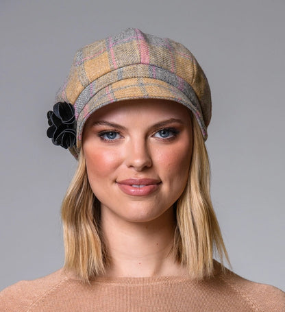 Frau mit schulterlangem, blondem Haar trägt eine karierte Schirmmütze mit schwarzer Blume und einen hellbraunen Pullover.