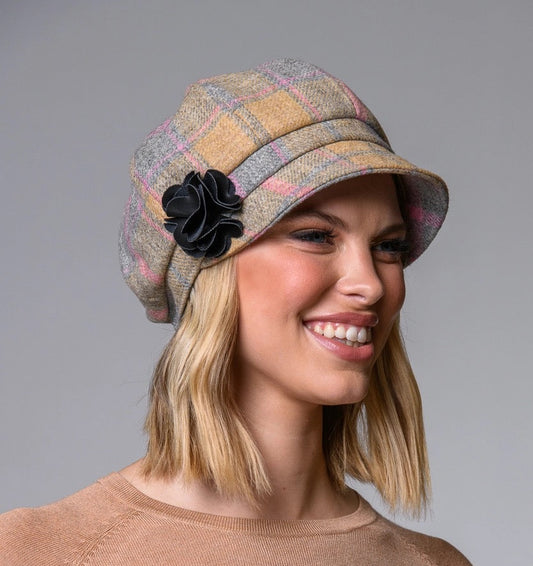 Frau mit schulterlangem, blondem Haar trägt eine karierte Schirmmütze mit schwarzer Blume und einen hellbraunen Pullover.