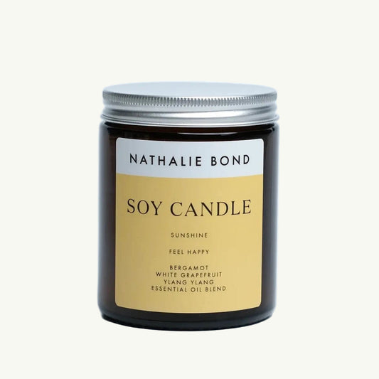 Braunes Glasglas mit silbernem Schraubdeckel, darauf ein gelb-beiger Aufkleber mit schwarzer Schrift 'NATHALIE BOND SOY CANDLE SUNSHINE FEEL HAPPY BERGAMOT WHITE GRAPEFRUIT YLANG YLANG ESSENTIAL OIL BLEND'. Duftkerze Sunshine